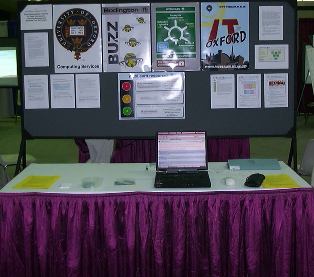 Educause 2006 poster. The board contains a variety of posters, promoting training courses and illustrating various uses of the WebLearn VLE, particularly personalisation with various symbology such as a roundabout with turn-offs for different uses. A laptop sits on the table, with a screen showing a demo CD.